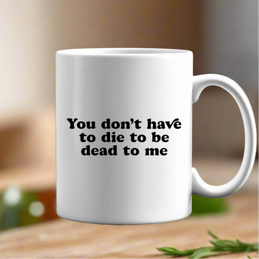The You Don’t Have To Die To Be Dead To Me Ceramic Mug by Kreative Kreationz features bold black text on white ceramic, displayed on a blurred wooden surface with greenery in the background.