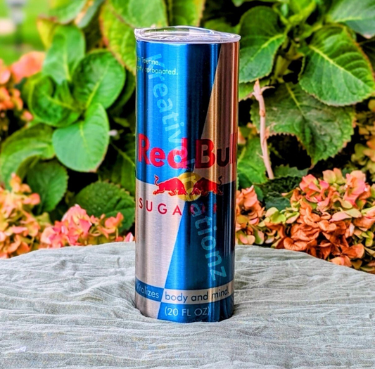 A Sugar Free Red Bull 20oz Tumbler by Kreative Kreationz stands upright on a gray cloth outdoors, with green leaves and pink hydrangea flowers in the background.