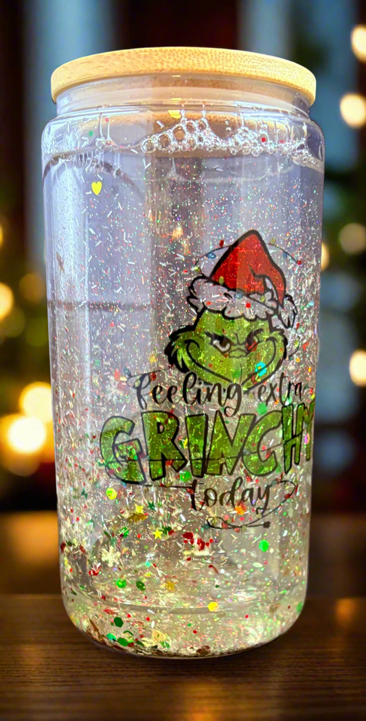 The Feeling Extra Grinchy Today Snowglobe Tumbler from Kreative Kreationz features a festive Grinch illustration with a Santa hat, filled with clear liquid and glitter. It's the perfect Christmas gift, enhanced by softly blurred warm lights.