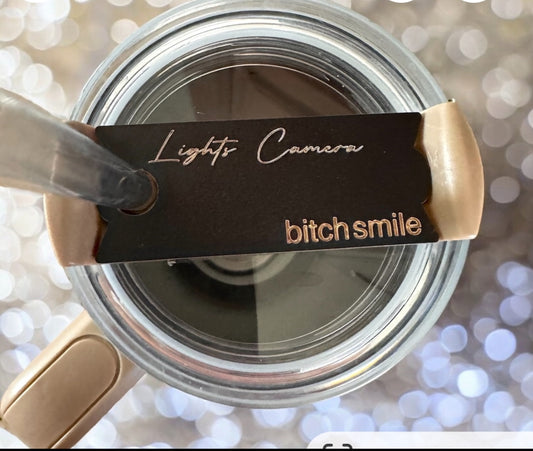 A close-up of an open cosmetic compact with a mirror resembles Kreative Kreationz's "I can do it with a broken heart" Stanley Lid Topper. The black lid displays "Lights Camera" in white and "bitch smile" in beige, while the background features bokeh light patterns for a personalized touch.