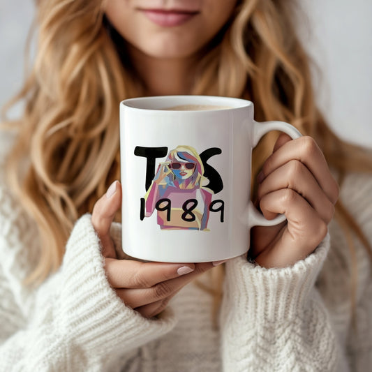 A person with long blonde hair and a white sweater holds the Taylor Swift 1989 Mug by Kreative Kreationz, featuring "T S," "1989," and an illustration of a woman in sunglasses. This mug captures the Swiftea Albums spirit, with the person's face partially visible.