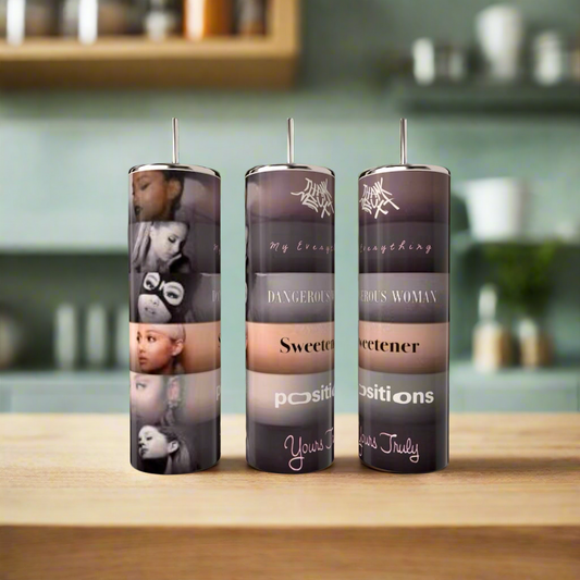 Three tall candles featuring Ariana Grande album art and song titles sit on a wooden surface beside the "Ariana Grande Album Aesthetic with External Sunshine 20oz Tumbler" by Kreative Kreationz in a modern kitchen with blurred shelves in the background.