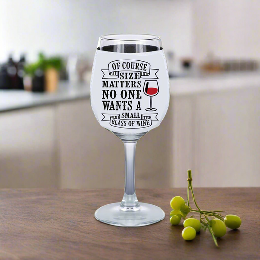 On a kitchen counter, a wine glass displays funny text about size: "Of course size matters, no one wants a small glass of wine." Surrounded by green grapes and set against a blurred kitchen background, it's ideal for true wine lovers who might fancy Kreative Kreationz's insulating neoprene Wine Glass Koozie.