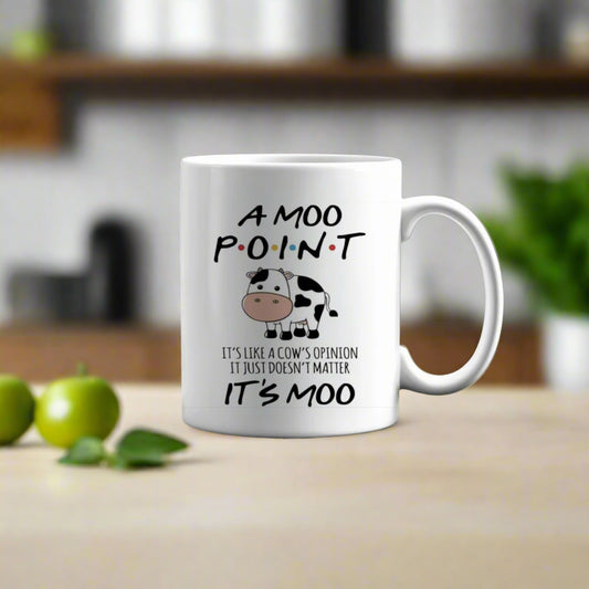 The Friends Moo Point Mug by Kreative Kreationz—a white ceramic mug with a cartoon cow and the quote, "A MOO POINT. It's like a cow's opinion, it just doesn't matter. It's moo"—rests on a mossy tree stump in a sunlit forest.