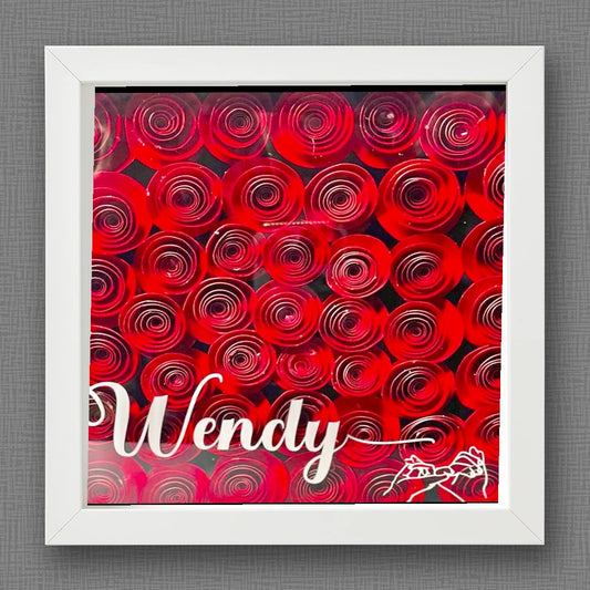 The Red Roses Flower Shadow Box | Personalized by Kreative Kreationz features tightly arranged red paper roses in a white frame, with "Wendy" and a rose illustration on the glass, set against a textured gray background—a unique keepsake.