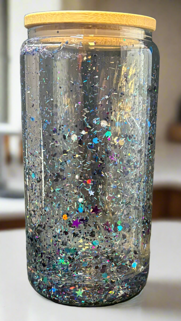 The Snowglobe Tumbler by Kreative Kreationz is a clear glass jar with a bamboo lid, filled with water and floating multicolored glitter, including silver stars—a charming handmade accent for any space.