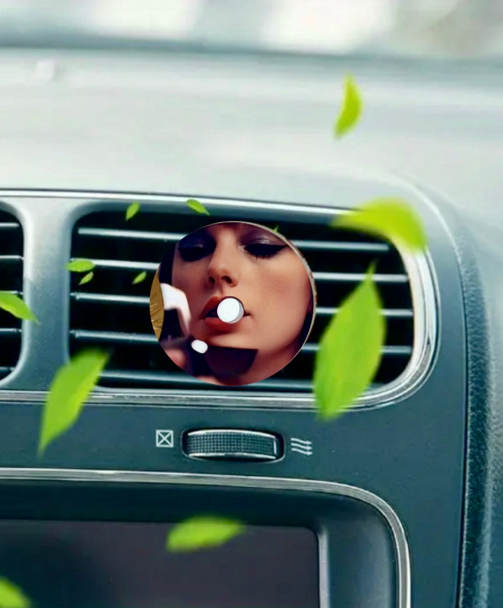 The Taylor Swift Car Air Freshener by Kreative Kreationz features a circular design of a woman touching up her lipstick, framed by green leaves—bringing fresh, stylish flair to your air vent.