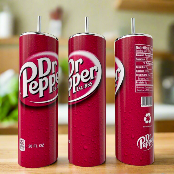 A Dr Pepper Stainless Steel Tumbler by Kreative Kreationz sits on a kitchen counter with three tall Dr Pepper cans nearby, showing the logo, nutrition facts, and recycling symbol against a blurred kitchen background.