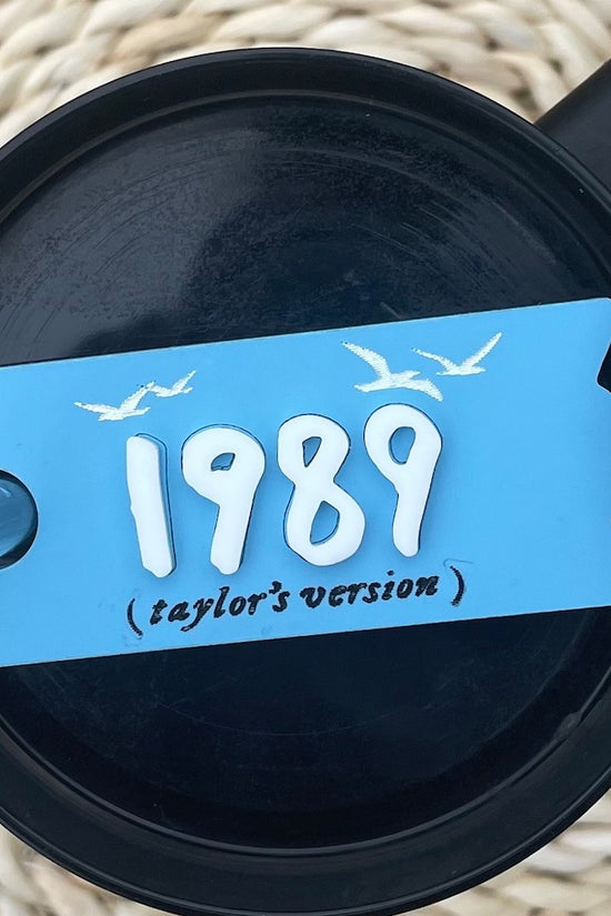 A clear, round plastic lid from a top-down view, featuring a gray sliding cover with a small circular opening on one side. The sliding cover has a logo of a winged lion wearing a crown. This customizable Kreative Kreationz Taylor Swift 1989 Taylor’s Version 20, 30, 40 ounce Stanley Lid Plate/Topper is perfect for adding flair to your drinkware collection.