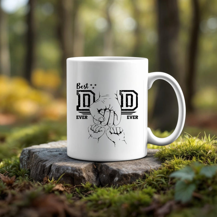 The Dad Best Ever Ceramic Mug by Kreative Kreationz, featuring two fists bumping and "Best Dad Ever," sits on a tree stump in a forest with vibrant green and yellow foliage blurred in the background. Perfect for coffee or tea lovers.