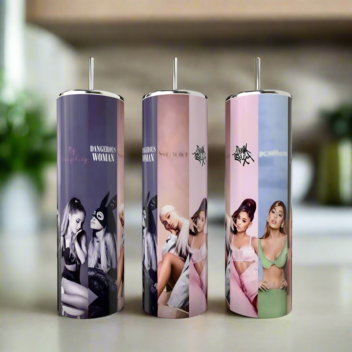 Three Kreative Kreationz Ariana Grande All Albums 20oz Tumblers, featuring images of Ariana in various poses and outfits, are placed on a table against a blurred indoor background.