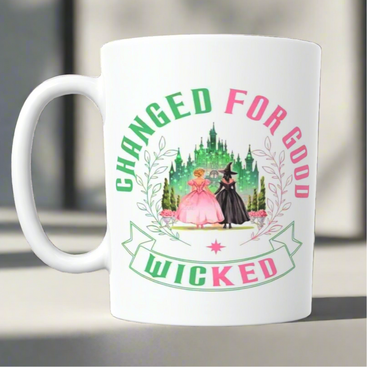 The Wicked Ceramic Mug by Kreative Kreationz showcases "CHANGED FOR GOOD" and "WICKED" with two women in pink and black dresses before a green castle, surrounded by decorative leaves.
