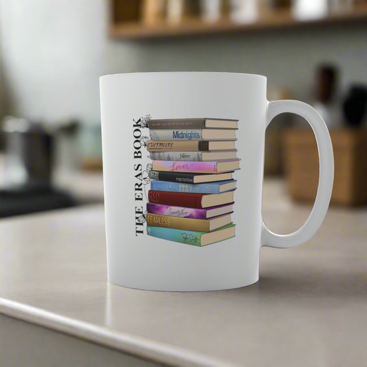 The Taylor Swift Eras Ceramic Mug by Kreative Kreationz features vibrant book stack designs titled "Midnights," "Red," and "Folklore" with "THE ERAS BOOK" text. Its 11oz ceramic form, set against a blurred kitchen background, makes it an ideal gift for any Swiftie.