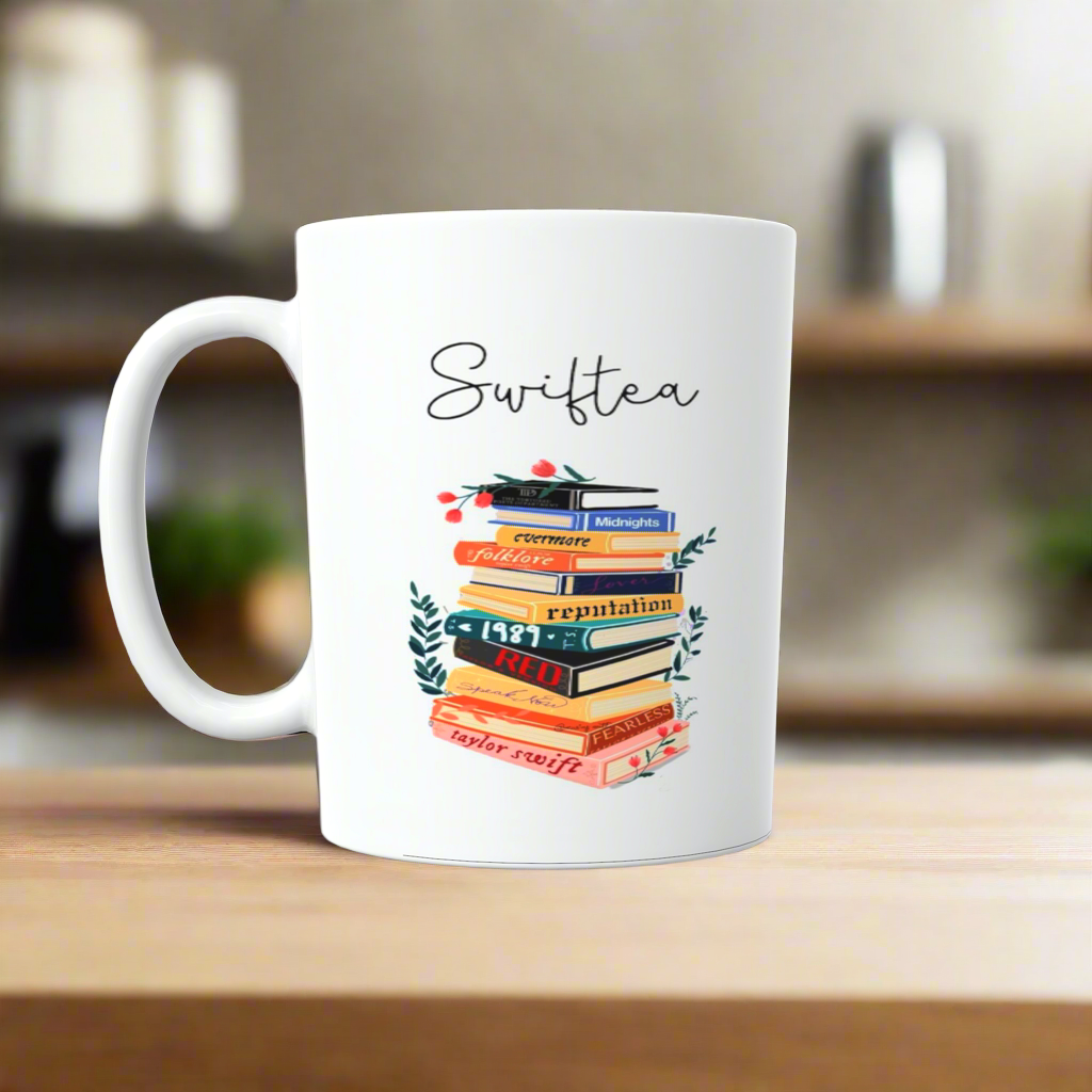 A white ceramic mug by Kreative Kreationz sits on a wooden surface featuring "Swiftea" above book-shaped illustrations labeled with album titles like "Lover" and "Folklore", each blooming with floral designs. This Swiftea Albums Mug-Taylor Swift Mug blends literary charm with musical homage.