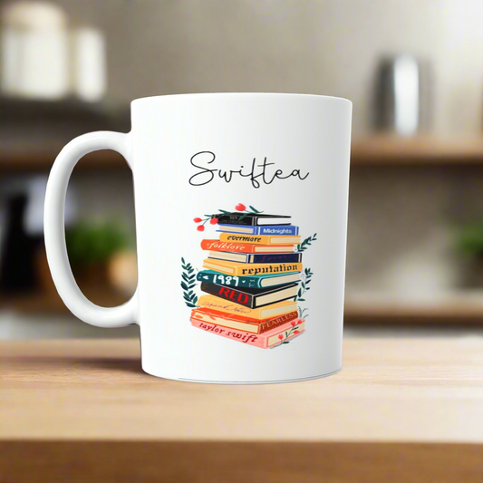 A white ceramic mug by Kreative Kreationz sits on a wooden surface featuring "Swiftea" above book-shaped illustrations labeled with album titles like "Lover" and "Folklore", each blooming with floral designs. This Swiftea Albums Mug-Taylor Swift Mug blends literary charm with musical homage.