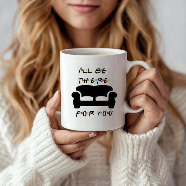 A woman with long, wavy hair holds a Kreative Kreationz Friends Couch Mug, a white ceramic piece decorated with "I'll be there for you" above an illustration of a black couch. She is dressed in a cozy white sweater.