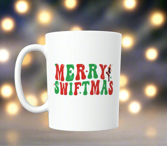 This merry season, enjoy your beverage in the Merry Swiftmas Taylor Swift Ceramic Mug by Kreative Kreationz, a white ceramic mug adorned with the text "MERRY SWIFTMAS" in festive red and green letters, featuring a small silhouetted figure of a woman. The backdrop showcases a cozy ambiance with warm, glowing lights.