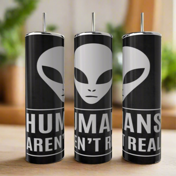 Three Kreative Kreationz Humans aren’t Real 20oz Skinny Tumblers feature a large white alien face above "HUMANS AREN’T REAL" on black stainless steel with silver lids and straws. They're eco-friendly and displayed on wood with a blurred green backdrop.