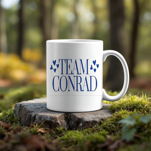 A "TEAM CONRAD" white mug with blue hearts rests on a tree stump in the woods—pair it with the Kreative Kreationz (Copy) Team Conrad Inspired Keychain for the perfect gift for fans.
