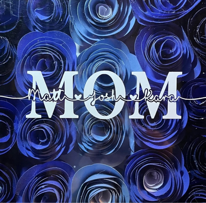 The Mom 10x10 Flower Shadow Box by Kreative Kreationz features "MOM" in bold white letters over blue and purple paper roses, with names Matt, Josh, and Kara above in cursive connected by a heart-shaped line.