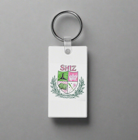 The Shitz University Keychain by Kreative Kreationz is a rectangular white accessory with a crest, green laurels, and shield icons—a book, castle, and pink flower—sublimation printed for vibrant detail.
