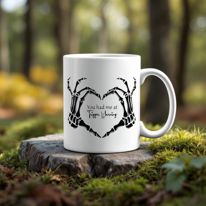 The Trigger Warning Mug by Kreative Kreationz, featuring skeleton hands forming a heart and "You had me at Tiffen Warning" text, rests on a tree stump against a blurred green forest backdrop—ideal for fans of unique photographic designs.