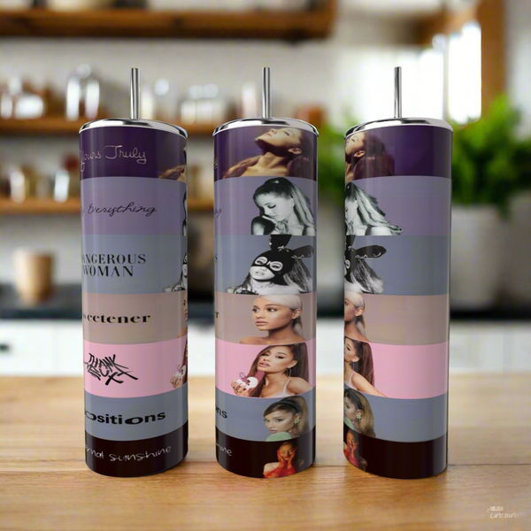 Three Kreative Kreationz Ariana Grande 20oz tumblers with metal straws sit on a kitchen counter, each decorated in vibrant horizontal bands featuring artistic portraits, song titles, and album covers.