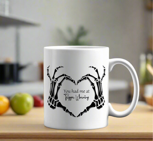 A white Kreative Kreationz Trigger Warning Mug sits on a kitchen counter, showing two skeleton hands forming a heart around the phrase "You had me at Trigger Warning," with blurred fruits and kitchen items in the background.