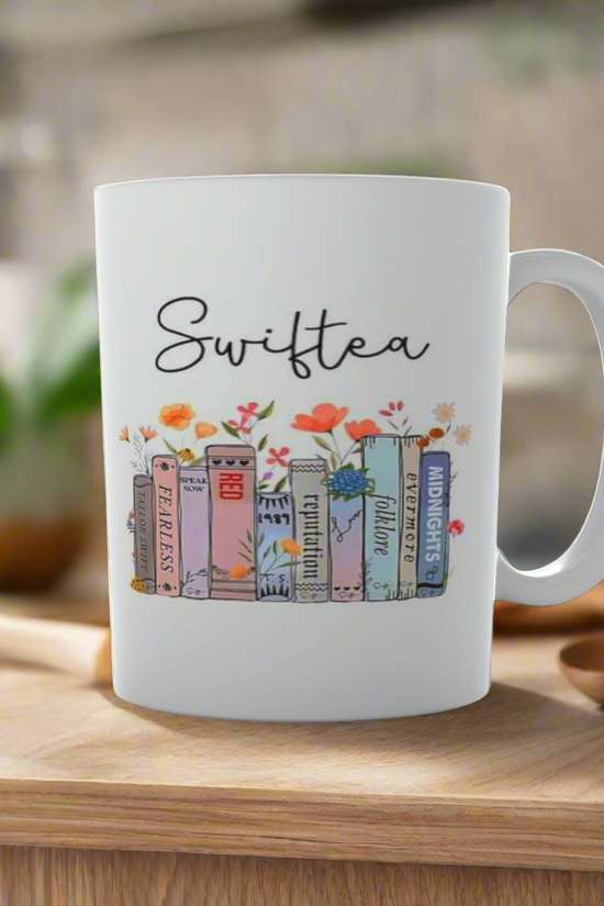 The Swiftea Mug-Taylor Swift by Kreative Kreationz is a white mug featuring vibrant book illustrations with album titles, embodying Taylor Swift's ERAS. It's set on wood against a blurred kitchen backdrop and small plant—ideal for fans seeking a cozy touch to their daily routine.