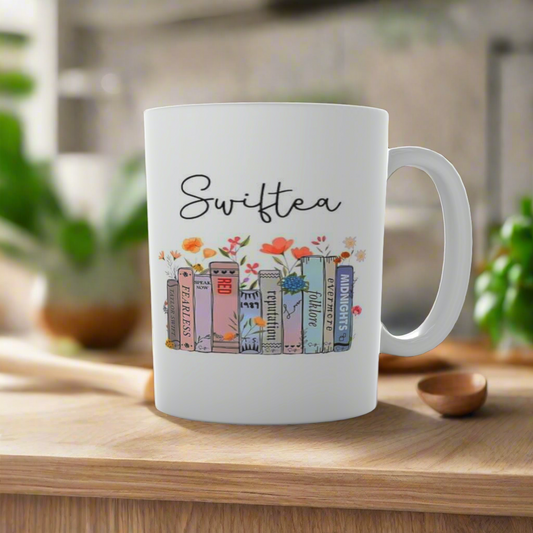 The Swiftea Mug-Taylor Swift by Kreative Kreationz is a white mug featuring vibrant book illustrations with album titles, embodying Taylor Swift's ERAS. It's set on wood against a blurred kitchen backdrop and small plant—ideal for fans seeking a cozy touch to their daily routine.