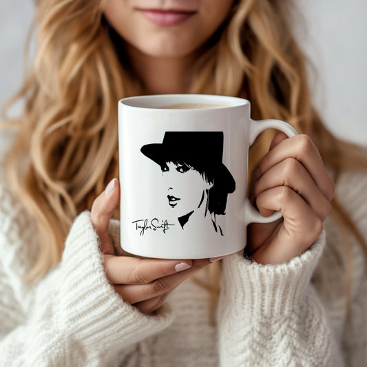 A person with long blonde hair, wearing a cozy white sweater, holds a Kreative Kreationz Taylor Swift Mug. The ceramic mug features a stylized illustration of a woman in a hat and signature-like text below the design.