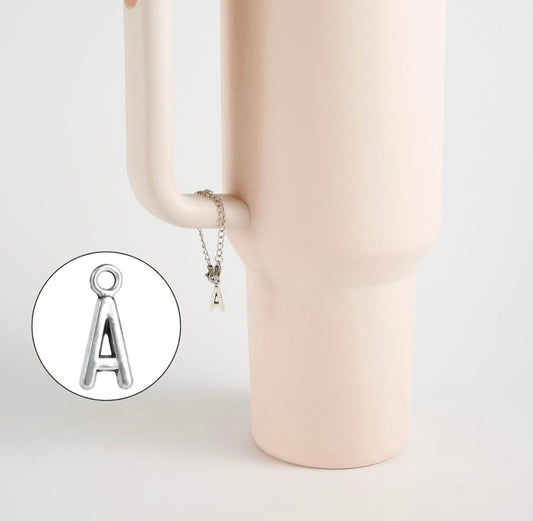A silver Letter Cup Charm Dangle by Kreative Kreationz hangs from the handle of a matte light pink vase. An inset highlights the "A" pendant’s detail, making it a stylish and unique drinkware accessory.