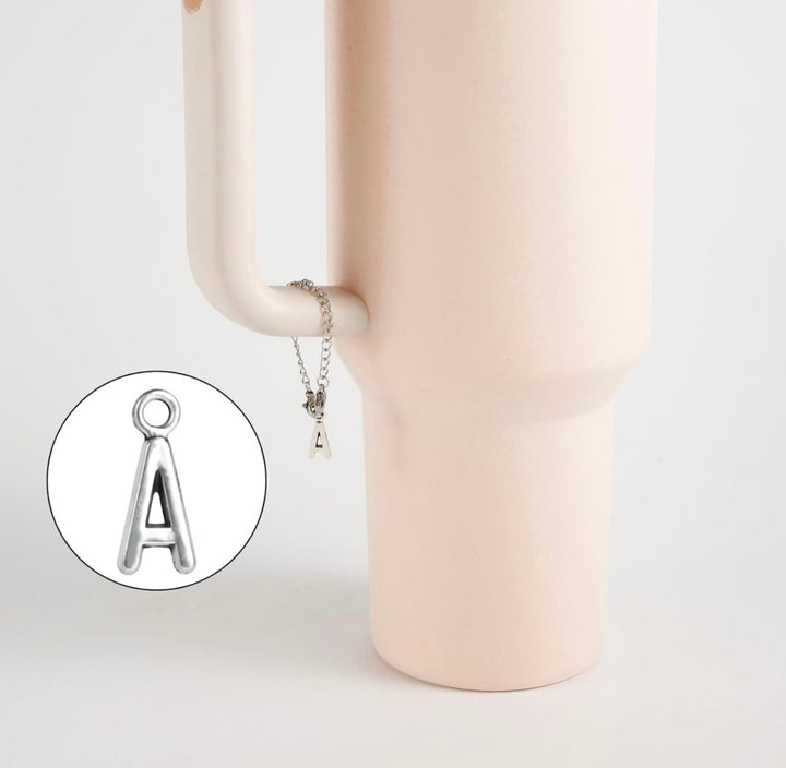 A silver Letter Cup Charm Dangle by Kreative Kreationz hangs from the handle of a matte light pink vase. An inset highlights the "A" pendant’s detail, making it a stylish and unique drinkware accessory.