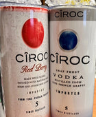 A lineup of CÎROC vodka cans in various flavors including original, mango, red berry, coconut, watermelon, and peach is beautifully arranged on an outdoor surface with a green hedge and blue sky background. Each can is paired with a chic 20oz Ciroc Tumbler with 3D Lid Topper by Kreative Kreationz.