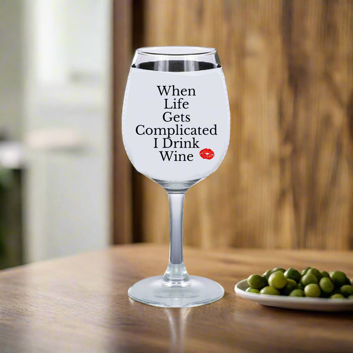 A wine glass with white liquid rests on wood, featuring "When Life Gets Complicated I Drink Wine" and a red lips design. Nearby is a dish of green olives and a Kreative Kreationz neoprene Wine Koozie, perfect for any wine enthusiast.