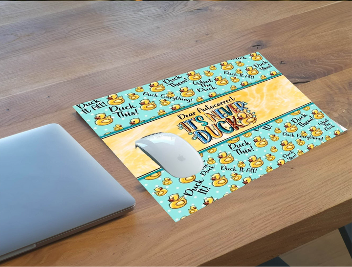 A laptop on a wooden table sits beside the Kreative Kreationz "Dear Autocorrect Its Never Duck Mousepad," featuring high-resolution cartoon rubber ducks and playful phrases.