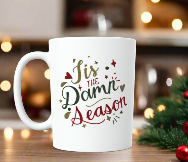 A "Tis The Damn Season Taylor Swift Ceramic Mug" from Kreative Kreationz rests on a wooden surface, set against a blurred festive background of warm lights and holiday decor. The white ceramic mug showcases the phrase "Tis the Damn Season" in a decorative red and green script, adorned with small festive graphics around the text.