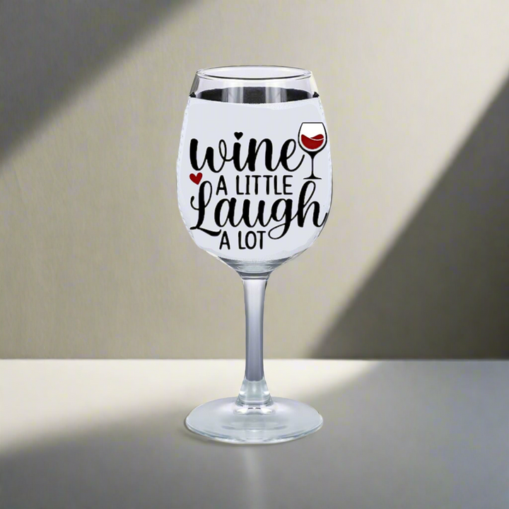 Kreative Kreationz's "Wne a Little Laugh Alot" wine glass, featuring a small red wine illustration, rests on a plain surface. Ideal for wine lovers, it pairs perfectly with the neoprene Wine Glass Koozie. The softly lit background enhances its inviting charm.
