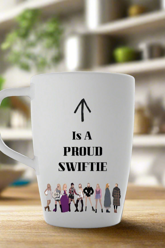 The Proud Swiftie Mug-Taylor Swift Mug by Kreative Kreationz features cartoon Taylor Swift illustrations from various eras, with “↑ Is A PROUD SWIFTIE” and an arrow toward the handle, against a blurred kitchen backdrop.