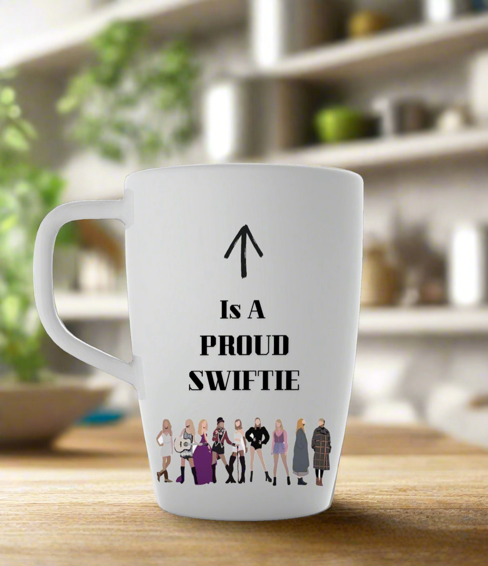 The Proud Swiftie Mug-Taylor Swift Mug by Kreative Kreationz features cartoon Taylor Swift illustrations from various eras, with “↑ Is A PROUD SWIFTIE” and an arrow toward the handle, against a blurred kitchen backdrop.