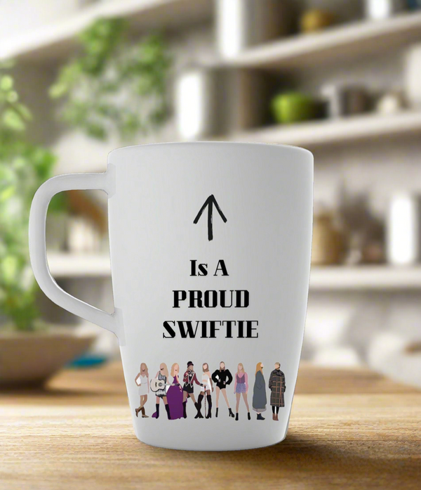 The Proud Swiftie Mug-Taylor Swift Mug by Kreative Kreationz features cartoon Taylor Swift illustrations from various eras, with “↑ Is A PROUD SWIFTIE” and an arrow toward the handle, against a blurred kitchen backdrop.
