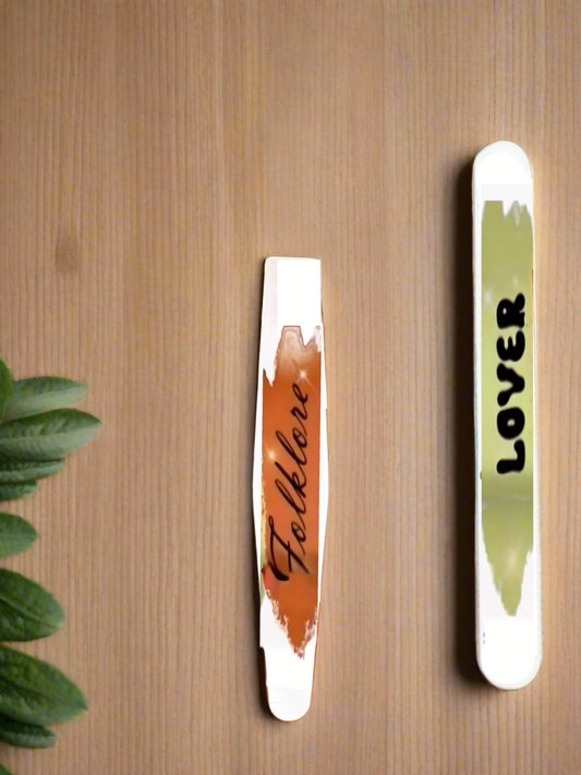 Two nail polish swatches, "Folklore" (brown) and "Lover" (light green), are shown on wood with green leaves—perfect Swifties nail art inspiration or to pair with your Kreative Kreationz Eras | 180 GRIT Stainless Steel Emery Board.