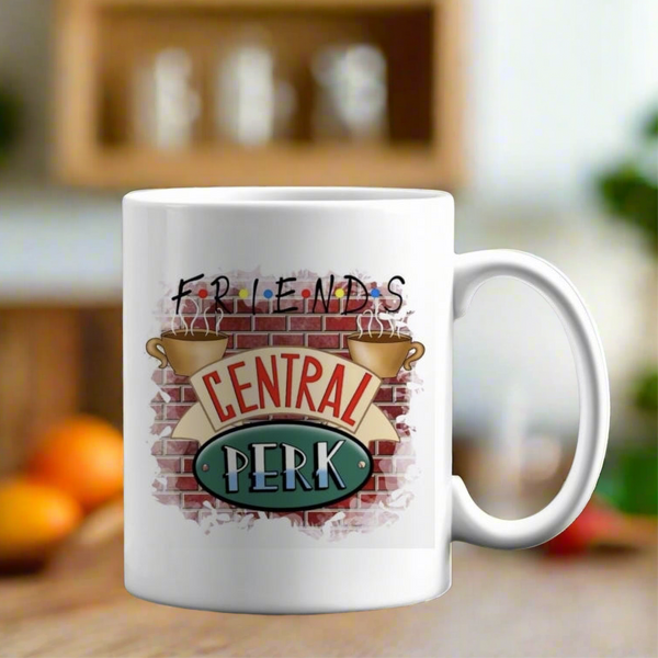 The Friends Central Perk Mug by Kreative Kreationz features the iconic TV show logo and Central Perk design with coffee cups and a brick wall motif, shown on a wooden surface against a blurred kitchen background.