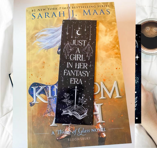 A copy of "Kingdom of Ash" by Sarah J. Maas rests on a bed, topped with Kreative Kreationz's "Just a girl in her fantasy era" bookmark, featuring stars, a moon, an open book, and floral designs.