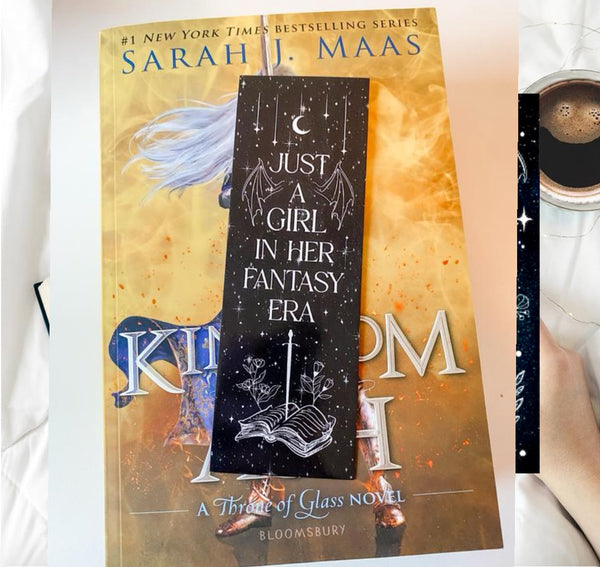 A copy of "Kingdom of Ash" by Sarah J. Maas rests on a bed, topped with Kreative Kreationz's "Just a girl in her fantasy era" bookmark, featuring stars, a moon, an open book, and floral designs.