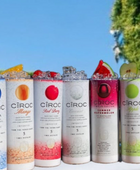 A lineup of CÎROC vodka cans in various flavors including original, mango, red berry, coconut, watermelon, and peach is beautifully arranged on an outdoor surface with a green hedge and blue sky background. Each can is paired with a chic 20oz Ciroc Tumbler with 3D Lid Topper by Kreative Kreationz.