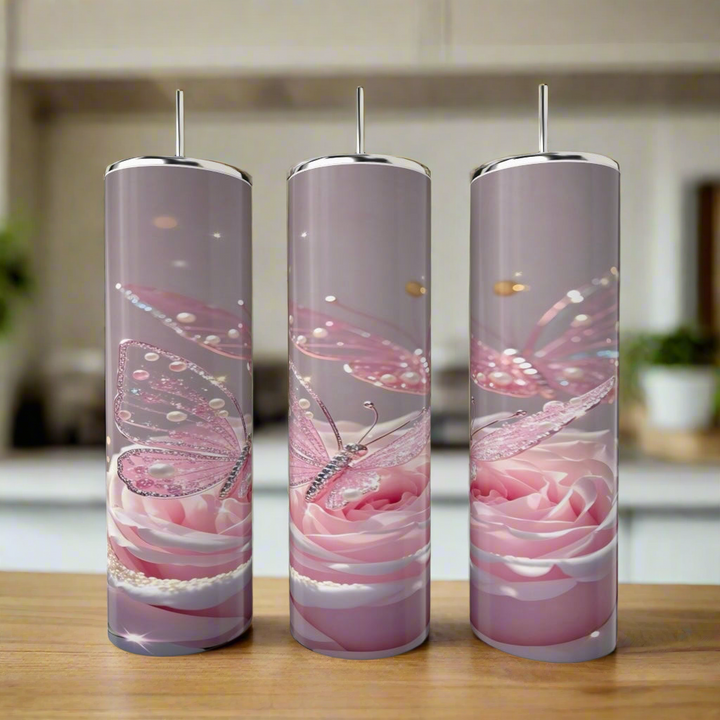 Three Kreative Kreationz Rose Butterfly 20oz Stainless Steel Tumblers with metal straws sit on a wooden surface. Each tumbler features pink butterflies and roses with sparkly accents on a soft lavender background. A kitchen is blurred behind.