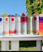 A lineup of CÎROC vodka cans in various flavors including original, mango, red berry, coconut, watermelon, and peach is beautifully arranged on an outdoor surface with a green hedge and blue sky background. Each can is paired with a chic 20oz Ciroc Tumbler with 3D Lid Topper by Kreative Kreationz.
