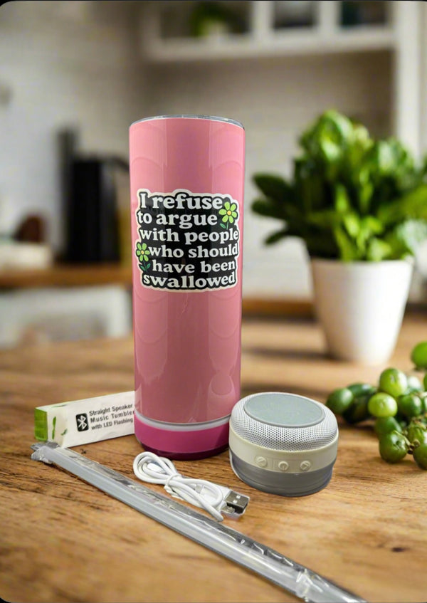 A Kreative Kreationz Customized 20oz Bluetooth Tumbler in pink, featuring "I refuse to argue with people who should have been swallowed," sits on a kitchen counter beside a portable speaker, metal straw, USB cable, and box. Fruit and plants are in the background.