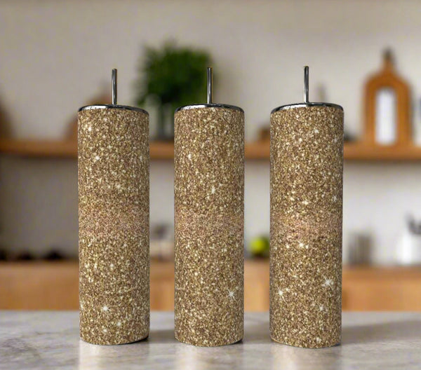 Three cylindrical spice shakers with brown, sand-like texture and silver tops are lined up on a kitchen counter next to the Gold Fearless Shimmer 20oz Skinny Tumbler by Kreative Kreationz, adding a touch of luxury drinkware to the scene.